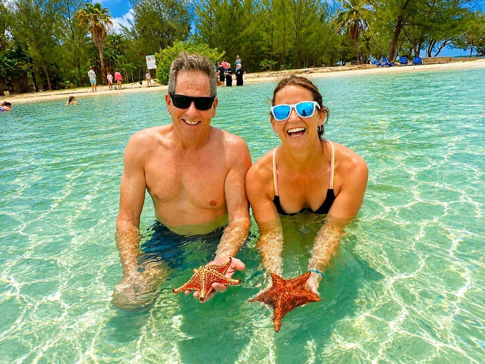 Laughing couple holding orange starfish in shallow turquoise water at Starfish Point on a Cayman Adventure Tours excursion in Grand Cayman