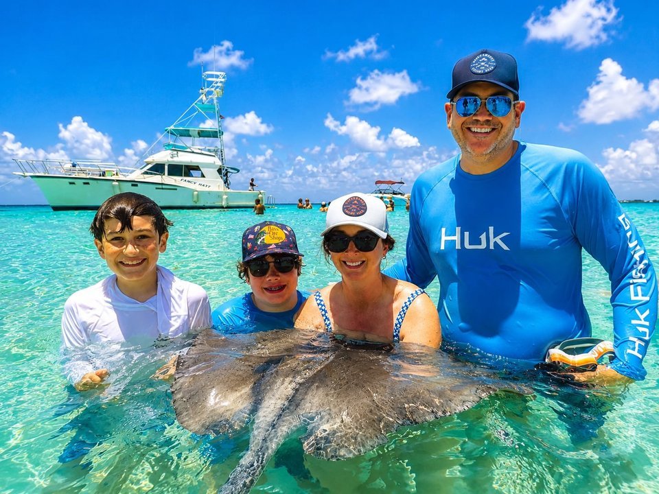 Family of four petting a large southern stingray at the Stingray City sandbar with the Cayman Adventure Tours boat anchored behind them in Grand Cayman
