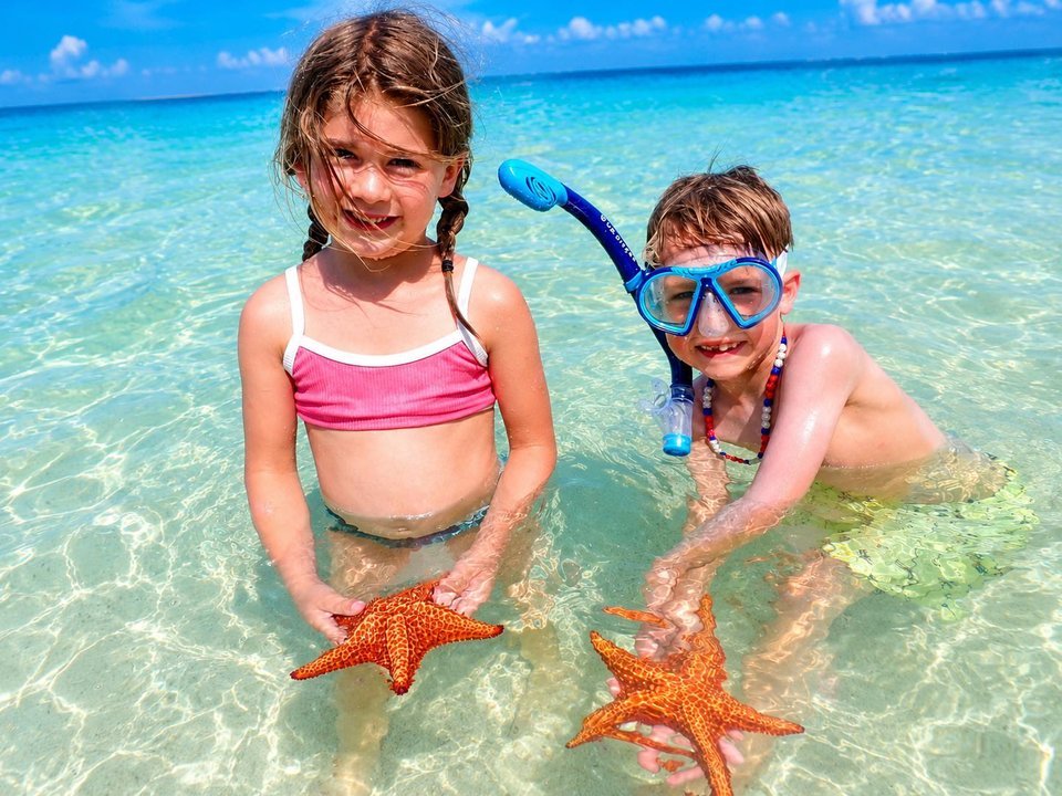 Two young children holding orange starfish in clear shallow water at Starfish Point during a Cayman Adventure Tours family trip in Grand Cayman